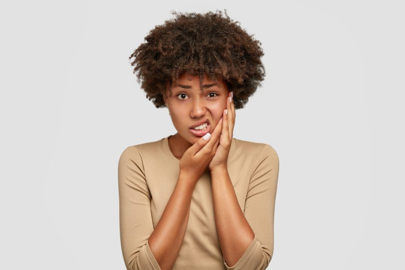 Isolated shot of displeased dark skinned young female has Afro haircut, keeps hand on cheek, frowns face from pain, suffers from toothache, dressed in casual sweater, expresses negative emotions